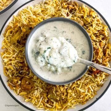 A spoon of biryani raita in a bowl along with spicy chicken biryani.