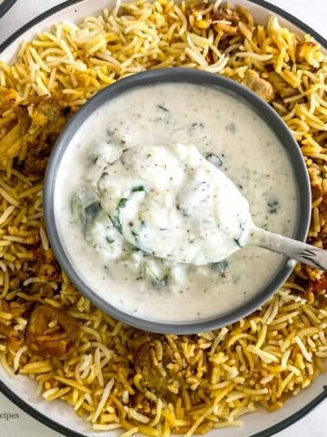A spoon of biryani raita in a bowl along with spicy chicken biryani.