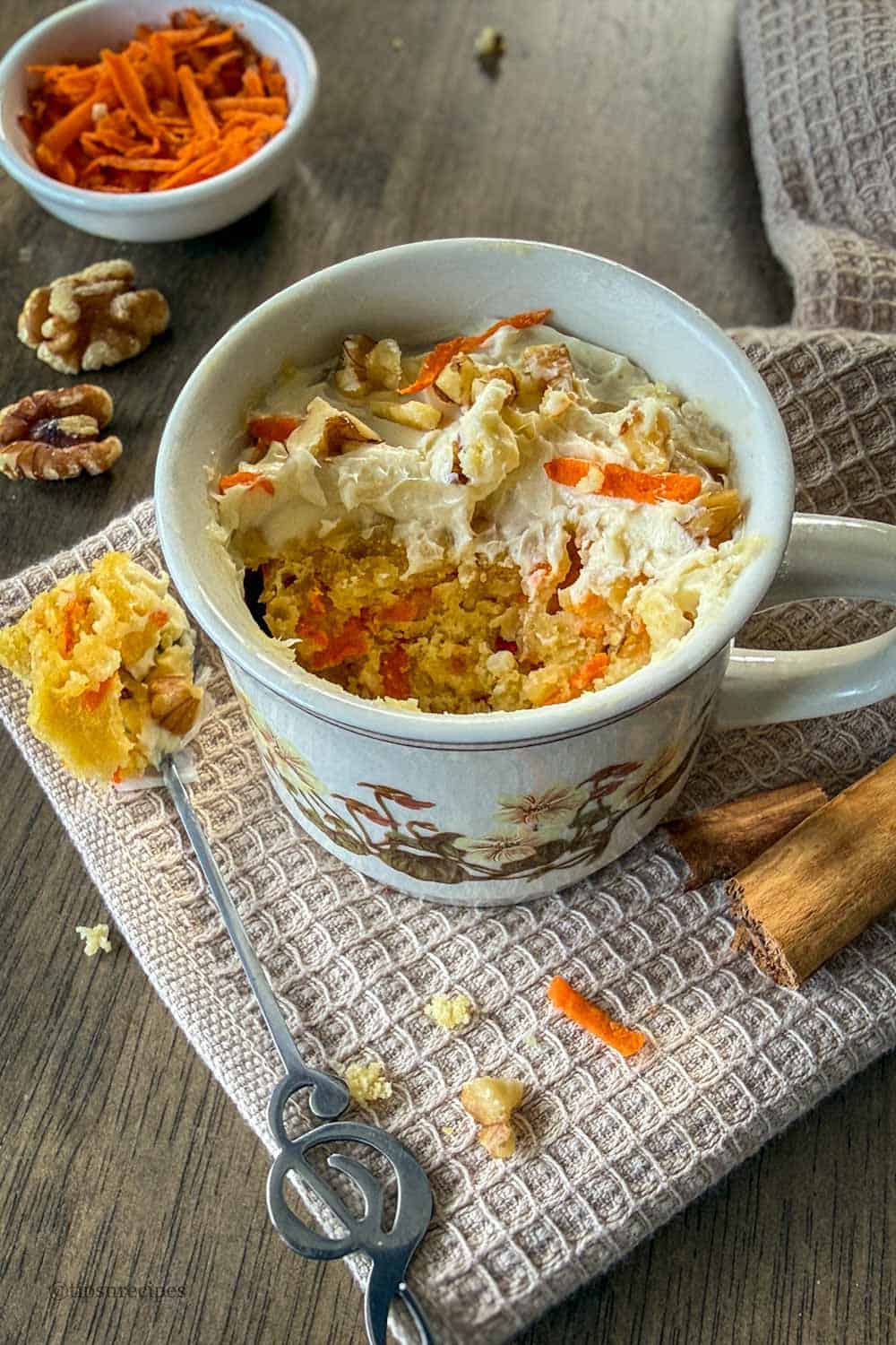 A mug cake of carrot cake with a bite in a spoon lying close to the mug.
